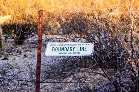 Image: against a backdrop of golden scrubland on a sunny day, a sign reading "United States Department of the Interior - Boundary Line - National Park service" hangs on a barbed wire fence.