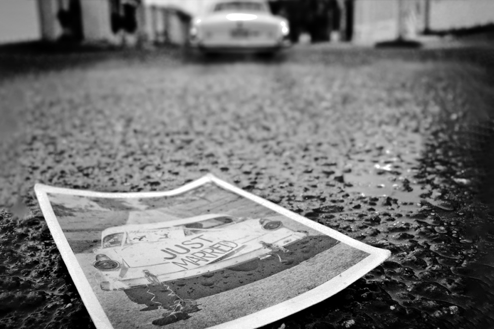Black and white photo of a rainy parking lot, with a single white car in the background. In the foreground is a discarded photo of that same car at some time in the past, when it was decorated with a sign reading 'JUST MARRIED.'