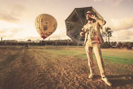 Image: on a large athletic field with a hot air balloon in the background, a man in fanciful Edwardian-style clothing stands with a parasol in one hand while he twirls the edge of his mustache with the other hand.