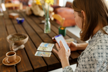 Image: on the dining table of a cheerfully-lit home with a cup of espresso nearby, a woman has spread three tarot cards while she consults her smartphone.
