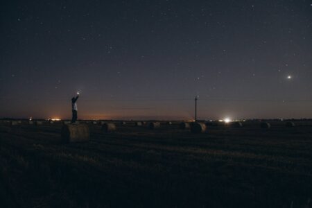 Image: on a dark night under a starry sky, a young man stands atop a hay bale holding aloft a small light as if trying to signal his presence to the universe.