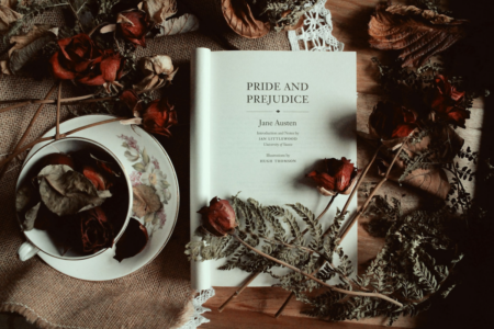 Image: a copy of Pride and Prejudice by Jane Austen open to the title page on a table surrounded by dried Autumn leaves and roses and a teacup.