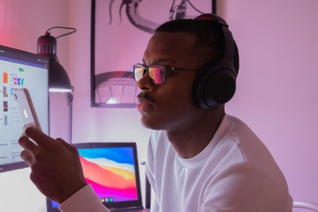 Image: a young man wearing headphones is staring at his smartphone while a desktop and laptop computer are also seen to be open in the background.