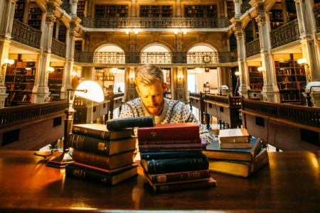 Image: In a large, richly detailed public library a man sits writing at a table, surrounded by stacks of reference books.