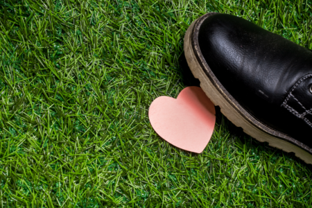 Image: a heart-shaped piece of pink construction paper lies on green grass where it's about to be stepped on by a man's black shoe.