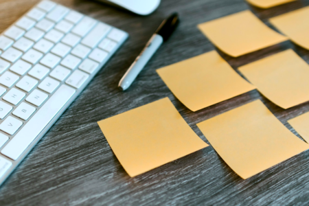 Image: Blank sticky notes are arranged in cascading rows (first one, then two, then three, then four) on a desktop next to a computer keyboard and black marker.