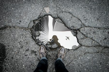 Image: A photographer stands at the edge of a puddle which has formed in broken blacktop after rain. He looks directly down at his feet and sees his own reflection in the puddle, as well as the reflection of a jet airplane passing overhead.