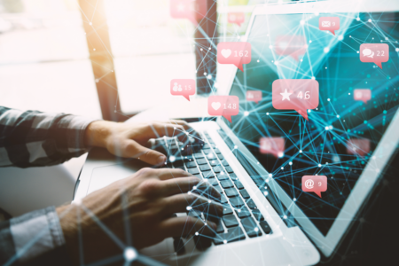 Image: a man types at a laptop computer, from which is bursting forth a constellation of implied networking connections and vectors of engagement such as alerts to new emails, mentions, reviews, messages, and likes.