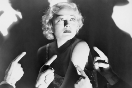 Image: black and white still from an early 20th century movie showing an elegant woman lit by a spotlight and wearing a concerned expression while numerous hands point fingers up at her in accusation.