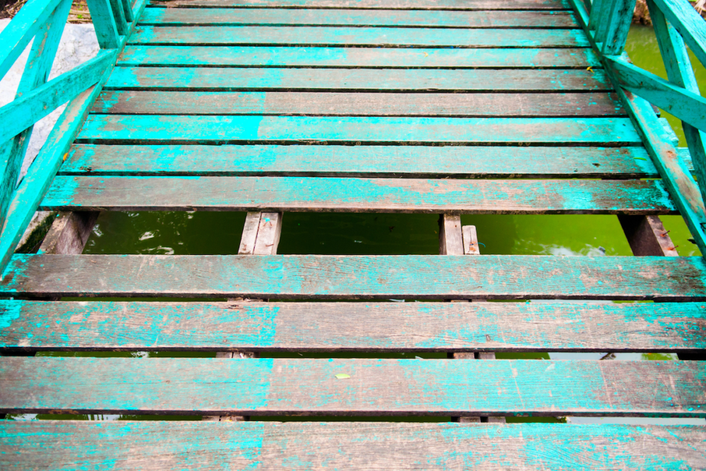 Image: a plank is missing from the floor of an old wooden bridge, revealing the green water beneath.