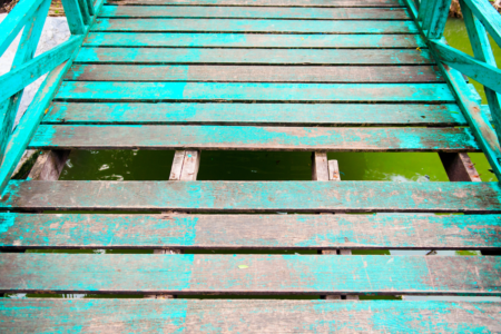 Image: a plank is missing from the floor of an old wooden bridge, revealing the green water beneath.