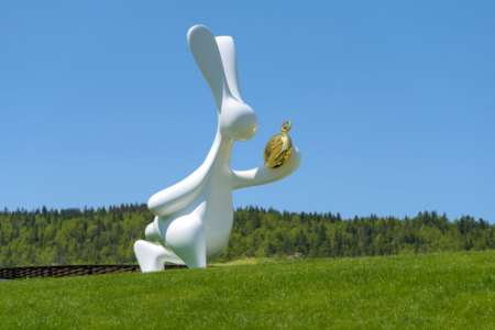 Image: modern sculpture of the rabbit from Alice's Adventures in Wonderland holding a gold watch, on the grounds of the Musée Atelier Audemars Piguet in Le Brassus, Switzerland.