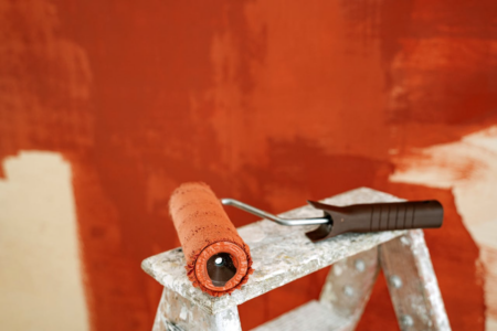 Image: against a backdrop of a wall in the process of being painted red, a paint roller sits atop a wooden stepladder.