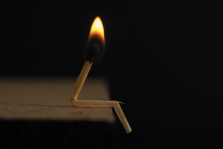 Image: in a darkened room, a lighted wooden match has been cracked and bent at two points so that it resembles a person sitting on the edge of a tabletop.