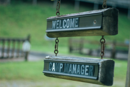 Image: a weathered wooden sign reading "Welcome Camp Manager" hangs from rusted chains against a backdrop of greenery and wooden fences.