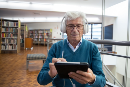 Image: In a spacious modern library is a senior man wearing headphones and interacting with a digital tablet device.