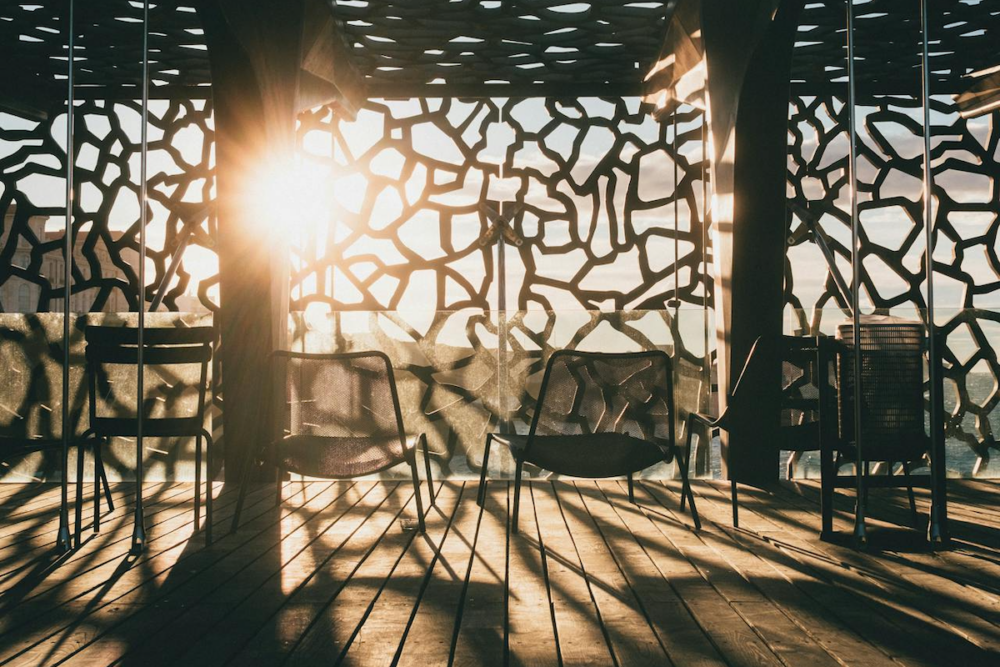 Image: Golden-hour sunlight filters through artistically-irregular cutouts of a floor-to-ceiling screen along the edge of an ocean-view patio where chairs are arranged and casting long shadows.