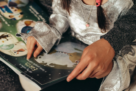 Image: a little girl sits on the lap of a woman, reading a picture book together.