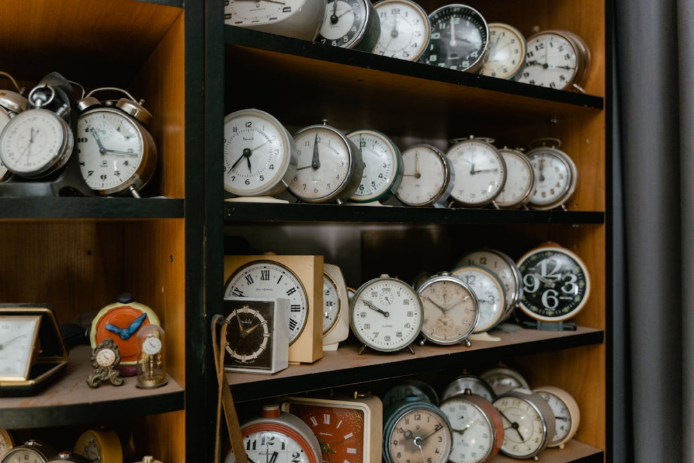 Image: rows of vintage alarm clocks and timers are arranged on wooden shelves.