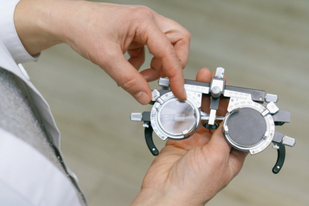 Image: an opthalmologist holds a diagnostic device shaped like a pair of eyeglasses outfitted with adjustable lenses.