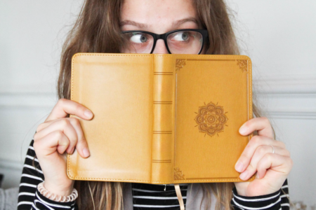 Image: a woman peers sidelong over the cover a book that she's holding up to hide her face.