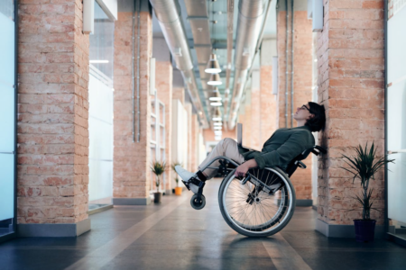 Image: a woman with a computer on her lap leans her wheelchair back against a brick column in a brightly lit modern office building.