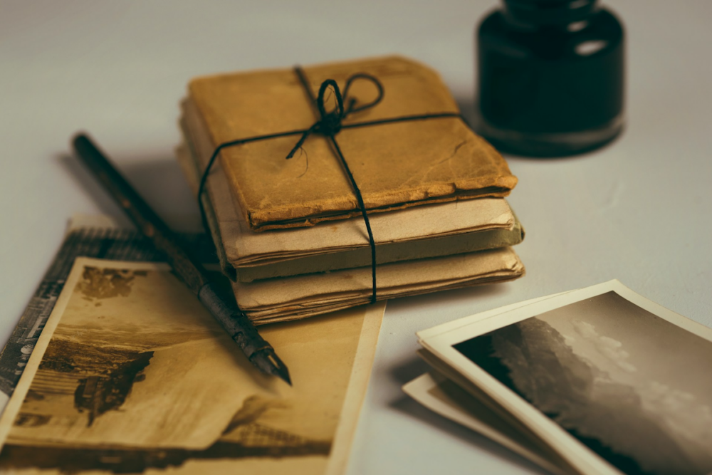 Image: a small stack of antique journals tied with string, alongside a fountain pen and some old black and white photographs.