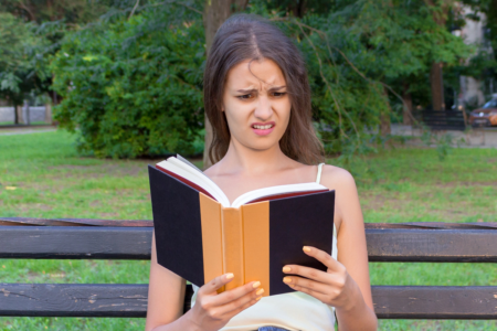 Image: a young woman sitting on a park bench wears a look of dismayed incredulity at something she's just read in the book she holds open in front of her.