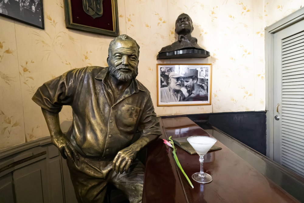 Image: a daiquiri cocktail and a freshly cut flower sit atop the bar next to the statue of Ernest Hemingway at El Floridita in Havana, Cuba.