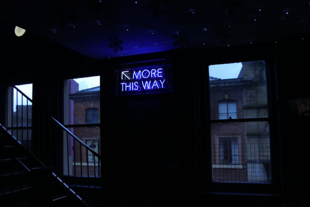 Image: in the darkened stairwell of an old urban high-rise building is a blue neon sign reading "MORE THIS WAY" with an arrow pointing up the stairs.