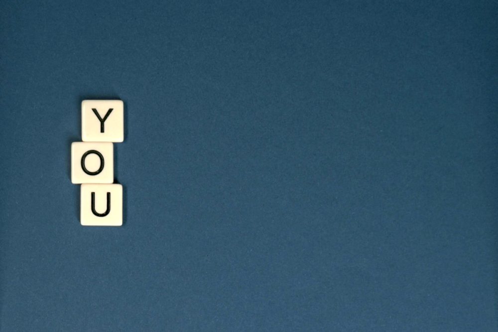 Image: Scrabble-style tiles of the letters Y-O-U on a blue background.