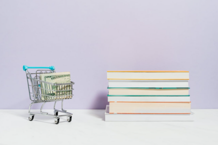 Image: next to a stack of books sits a miniature shopping cart with a folded $20 bill inside.