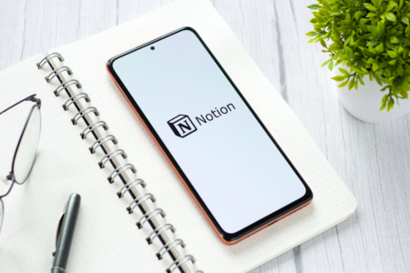 Image: A smartphone displaying the Notion app logo sits atop an open spiral-bound notepad alongside a pen, a pair of eyeglasses, and a small potted plant.