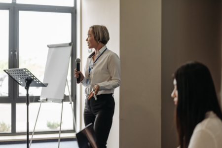 Image: a woman stands with a microphone in one hand and a marker in the other, prepared to make notes on a large pad of paper while giving a presentation to an audience in a well-lit modern office-style room.