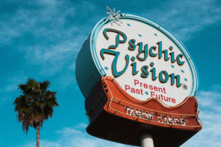 Image: in Los Angeles, against a blue sky and near a palm tree, is a towering neon sign advertising "Psychic Vision: Present, Past, Future • Tarot Cards".