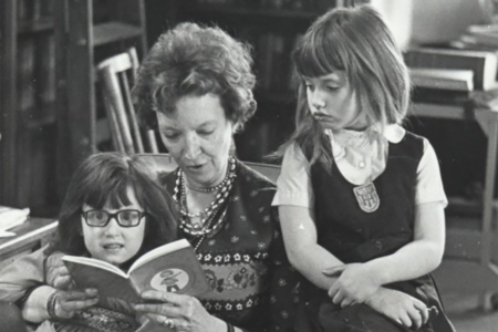 Image: black and white photo of the author Madeleine L'Engle sitting in a chair and reading to her granddaughters Charlotte, who sits on L'Engle's lap, and Lena who sits on the chair's arm.