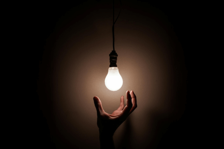 Image: in a darkened space, a hand reaches up from below to snatch a single glowing lightbulb hanging from an electrical wire.