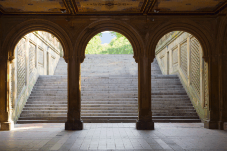 Image: the view from within Bethesda Terrace Arches, Central Park, New York, standing within a darkened space looking out at a grand staircase leading up into a brightly-lit day.