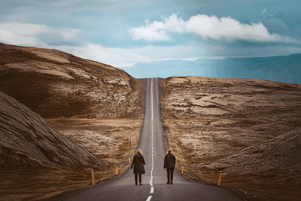 Image: two women walk together on either side of the painted lines along a remote road that leads up a steep hill near Reykjavik, Iceland.