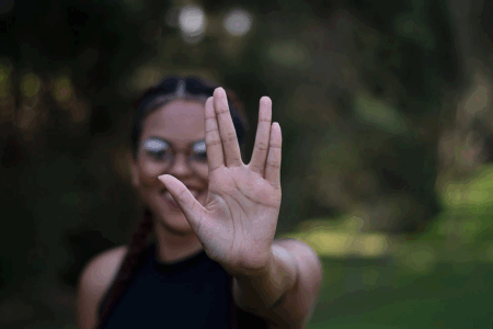 Image: a young woman standing in the outdoors gives the Vulcan hand salute to the viewer