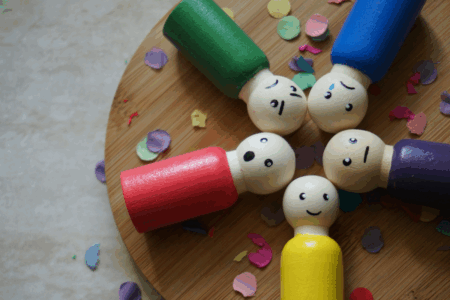 Image: five small cylindrical wooden figurative toys with painted faces representing emotions of happiness, surprise, anger, sadness, and neutrality are arranged on a wooden board with confetti strewn around them.
