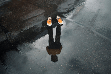 Image: a pair of unoccupied shoes sits at the edge of a puddle on a sidewalk. In the puddle is seen the reflection of the person who ought to be occupying the shoes.