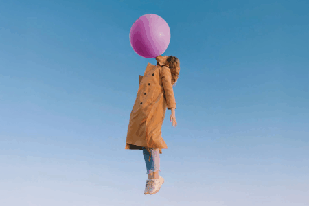 Image: a woman wearing jeans, athletic shoes and an open raincoat is carried into the air by the enormous pink bubble she's blown with chewing gum.