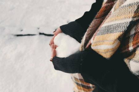 Image: Against a backdrop of snowy ground, a woman wearing a sweater and scarf holds in her clasped hands a heart-shaped mound of snow.
