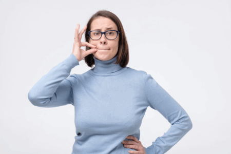 Image: a brown-haired woman wearing glasses and a light blue turtleneck sweater holds one hand to her mouth in a "zipped lips" gesture.