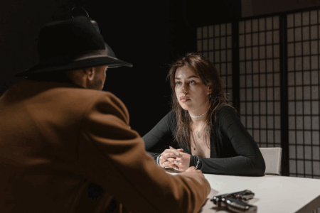 Image: a young woman wearing handcuffs sits at a table, staring blankly at the man sitting across from her who has a gun at his side.