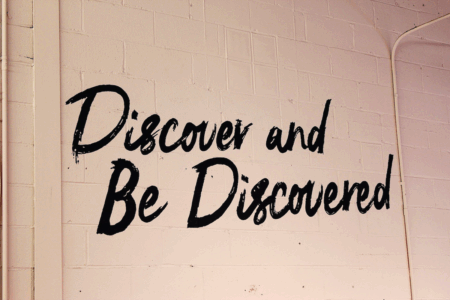Image: The words "Discover and Be Discovered" are painted in cursive style on a light pink cinderblock wall.