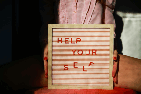 Image: a seated woman holds a small sign with adjustable letters that are arranged to spell the message "Help your self".