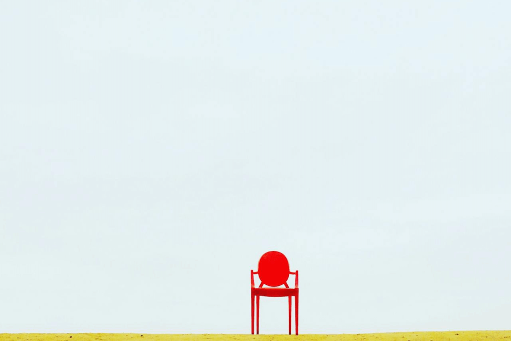 Image: on a thin strip of green ground, a bright red chair stands alone against a vast expanse of blue sky.