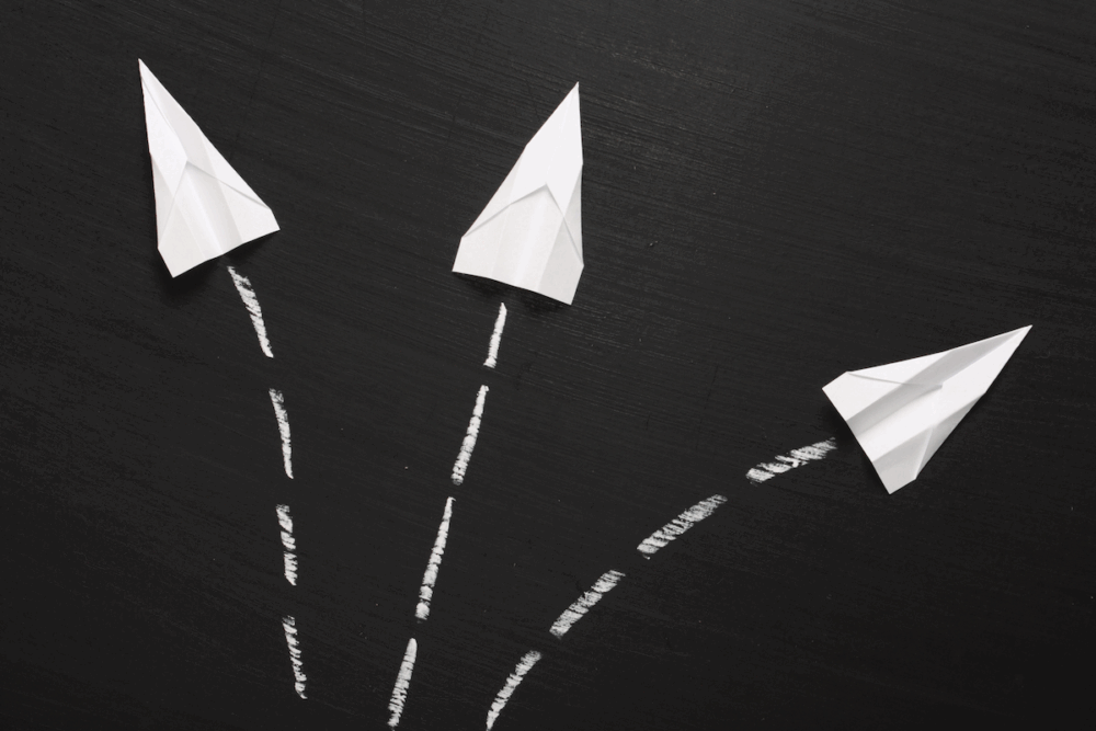 Image: three white paper airplanes lie atop a blackboard, aimed in different directions with white chalk marks behind them indicating their paths.